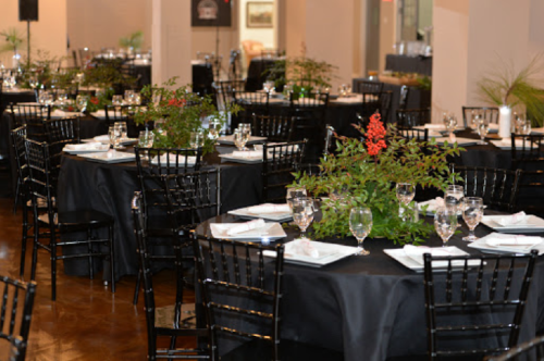 Elegant dining setup with black tablecloths, glassware, and centerpieces of greenery in a spacious venue.