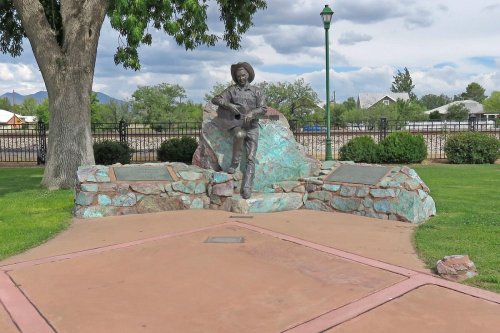 A bronze statue of a musician playing guitar, set on a stone base in a park with green grass and trees.