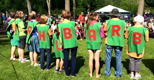 A group of people wearing green capes with red letters spelling "Rhubarb" at an outdoor event.