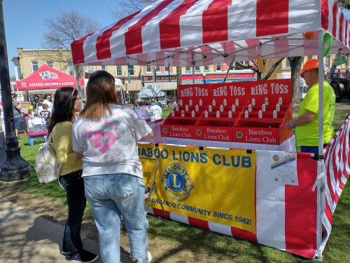 A colorful ring toss game booth at a fair, with two people interacting with a vendor from the Baraboo Lions Club.