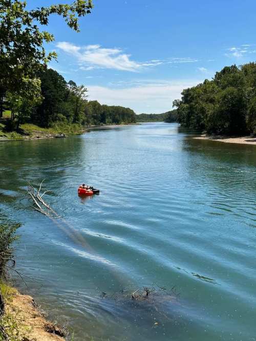 A calm river scene with a person in an orange float, surrounded by lush greenery and a clear blue sky.
