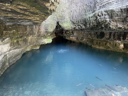 A serene cave with a clear blue pool of water, surrounded by rocky walls and a dark entrance in the background.