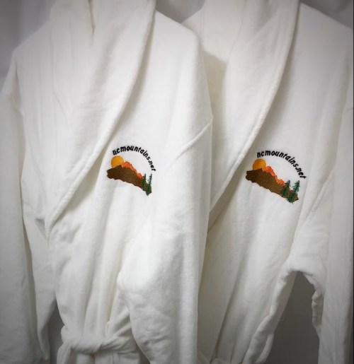 Two white bathrobes hanging side by side, each featuring an embroidered mountain design.