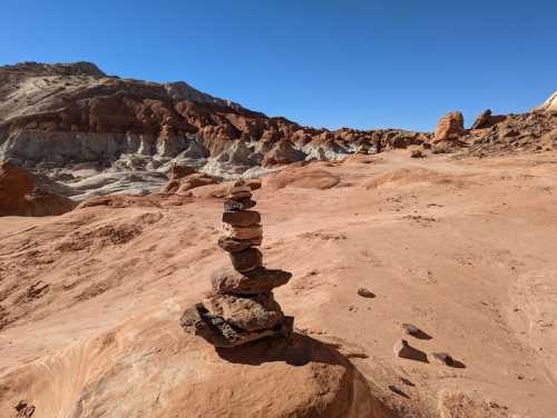 A stack of rocks balanced on a sandy desert landscape with colorful rock formations in the background under a clear blue sky.