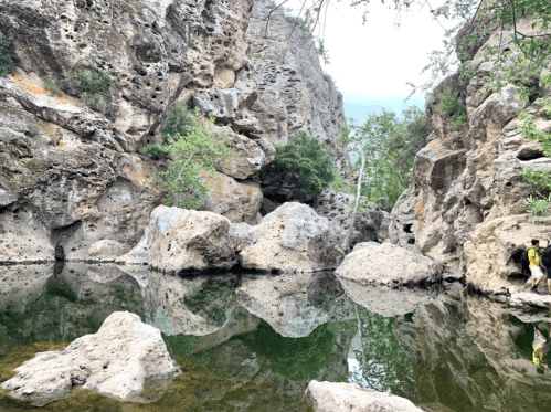 A serene rocky landscape with a calm water pool reflecting the surrounding cliffs and greenery.