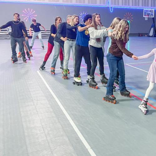 A group of people roller skating in a line at a rink, with colorful lights in the background.