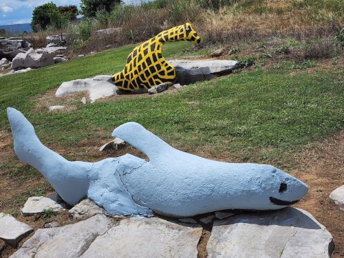 A blue shark sculpture and a yellow giraffe sculpture on a grassy area with rocks.