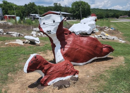 A large, painted rock sculpture of a fox and a smaller fox, set in a grassy area with trees in the background.