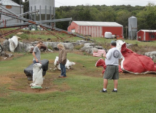 Children exploring a farm with large, colorful sculptures in a grassy area, surrounded by barns and silos.