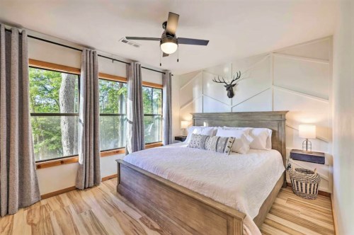 Cozy bedroom with a large bed, wooden accents, and windows overlooking trees, featuring a ceiling fan and decorative antlers.