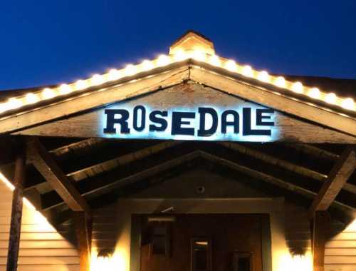 A building entrance at night with a sign reading "ROSEDALE" illuminated by lights on the roof.