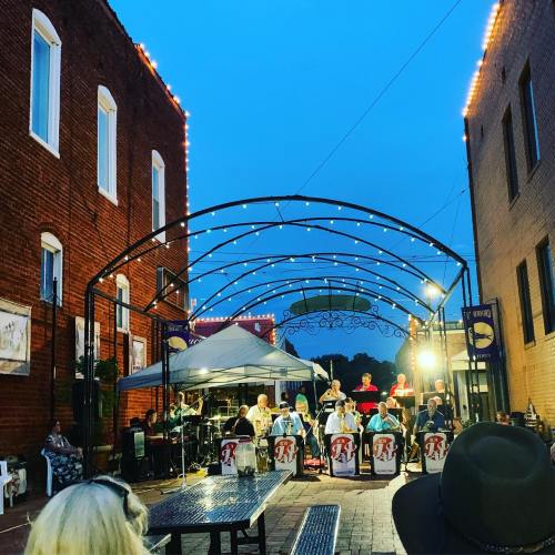 A jazz band performs under string lights in a brick alleyway during twilight, with an audience seated nearby.