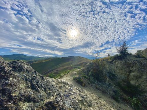 A rocky landscape under a cloudy sky, with the sun shining brightly in the center and rolling hills in the background.