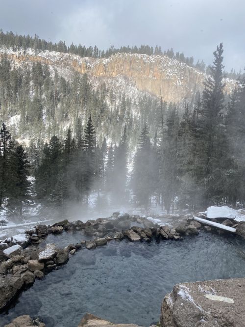 A steaming hot spring surrounded by rocky terrain and snow-covered trees, with a misty mountain backdrop.