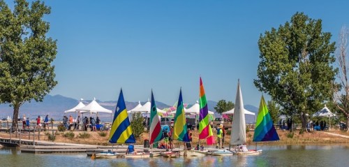Colorful sailboats on a calm lake with people and tents in the background under a clear blue sky.