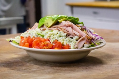 A colorful salad in a bowl featuring lettuce, tomatoes, avocado, ham, and blue cheese on a wooden surface.
