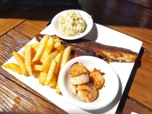 A plate with seasoned fish, scallops, crinkle-cut fries, and a side of coleslaw on a wooden table.