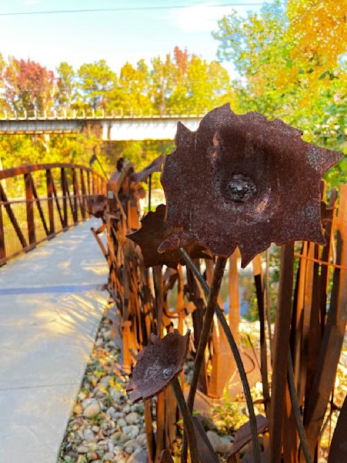 A pathway lined with metal flower sculptures, surrounded by colorful autumn foliage and a bridge in the background.