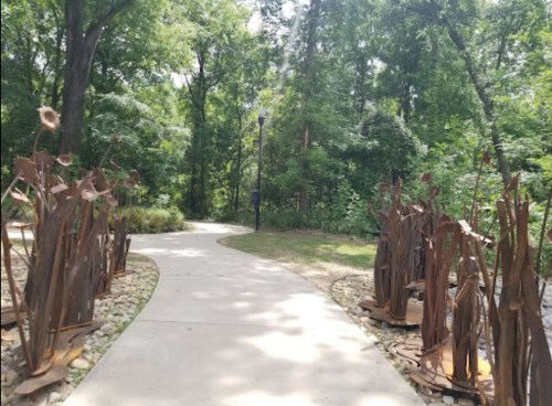 A winding path through a lush green forest, flanked by artistic metal sculptures and natural landscaping.