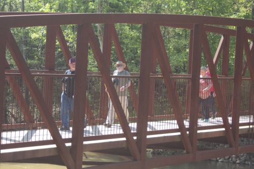 Three people walking on a metal bridge surrounded by greenery on a sunny day.