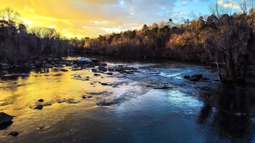 A serene river at sunset, reflecting golden and blue hues, surrounded by trees and rocky banks.
