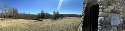 A panoramic view of a grassy landscape with trees and a stone structure on the right under a clear blue sky.