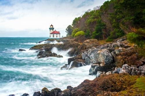 A lighthouse stands on a rocky shore, surrounded by trees, with waves crashing against the rocks under a cloudy sky.