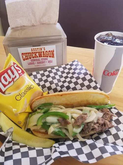 A sandwich with steak, peppers, and onions, served with chips, a pickle, and drinks in a casual dining setting.