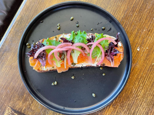 A black plate with a slice of bread topped with smoked salmon, greens, pickled onions, and capers.