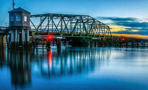 A green metal bridge spans a calm waterway at sunset, reflecting colorful lights and clouds in the water.
