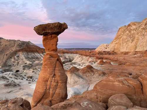 A towering rock formation with a flat top stands amidst colorful desert landscapes under a pastel sky at sunset.