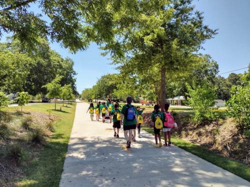 A group of children in green shirts and backpacks walking along a sunny path lined with trees.