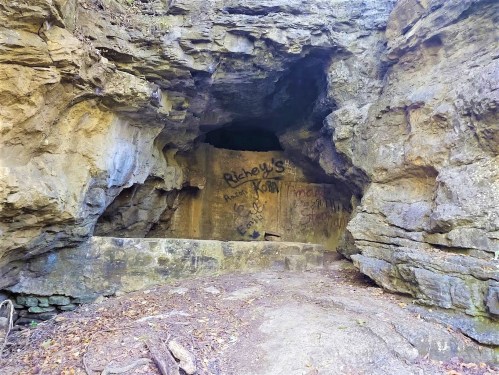A rocky cave entrance with graffiti on the walls, surrounded by natural stone formations and forest debris.