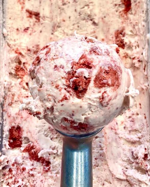 A close-up of a scoop of red velvet ice cream on a metal scoop, with swirls of cream and cake crumbs in the background.