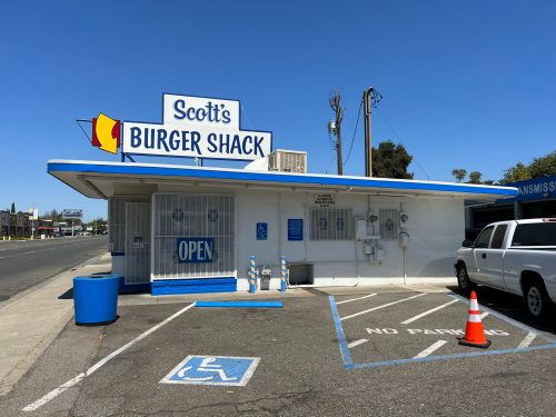 A small fast-food restaurant, Scott's Burger Shack, with a blue and white exterior, open sign, and parking restrictions.