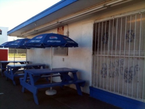 Blue picnic tables with Pepsi umbrellas outside a building with a barred window and a white wall.