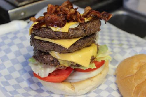 A towering burger with three beef patties, melted cheese, lettuce, tomato, and crispy bacon on top, served on a bun.