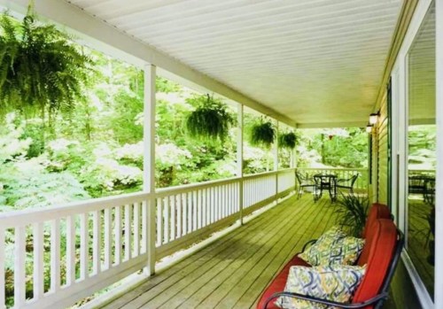 A serene porch with a red bench, potted plants, and hanging ferns, surrounded by lush greenery.