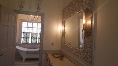 A stylish bathroom entrance featuring a vintage mirror, wall sconces, and a stained glass window above a freestanding tub.