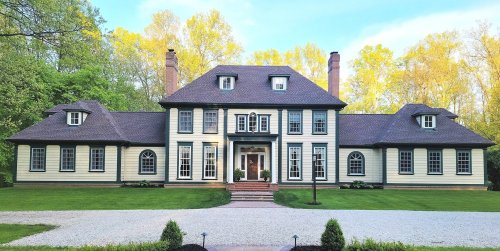 A large, elegant two-story house with a symmetrical facade, surrounded by greenery and a gravel driveway.