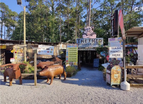 Entrance to "The Shed" with wooden pig sculptures, colorful signs, and a gravel path surrounded by trees.