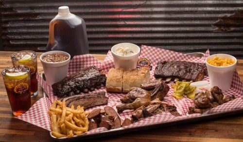 A large platter of assorted barbecue meats, sides, and drinks, served on a checkered paper-lined tray.
