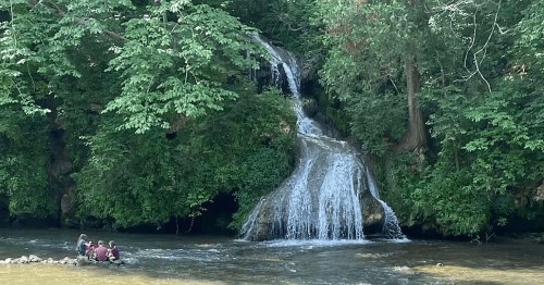 A serene waterfall surrounded by lush greenery, with people sitting by the water's edge.