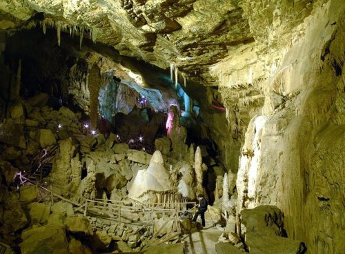 A dimly lit cave with stalactites and stalagmites, featuring colorful lights and a wooden walkway for exploration.