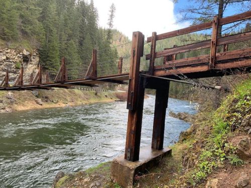 A wooden suspension bridge spans a river, surrounded by lush green trees and rocky terrain.