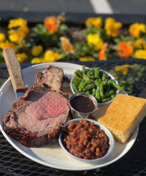 A plate featuring a juicy prime rib, green beans, baked beans, cornbread, and a side of sauce, with flowers in the background.