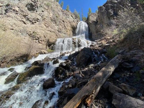 A cascading waterfall flows down rocky terrain, surrounded by trees and sunlight in a natural landscape.