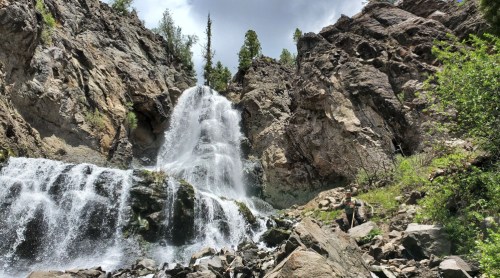 A cascading waterfall flows over rocky cliffs, surrounded by greenery and trees under a cloudy sky.