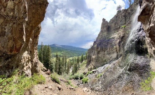 A panoramic view of a rocky canyon with lush greenery and a waterfall under a cloudy sky.