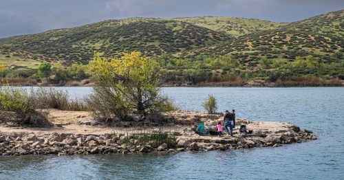 Father and small kids setting up fishing spot on rocky shoreline pointing in blueish water under heavy cloudscape. Green hills on horizon.